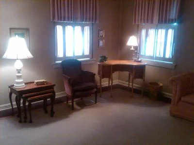 Quiet living room with lamp, chairs, desk, and bright windows behind curtains