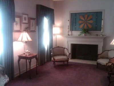 Cozy living room with purple carpet, chairs, lamps, and a fireplace with colorful wall art above it