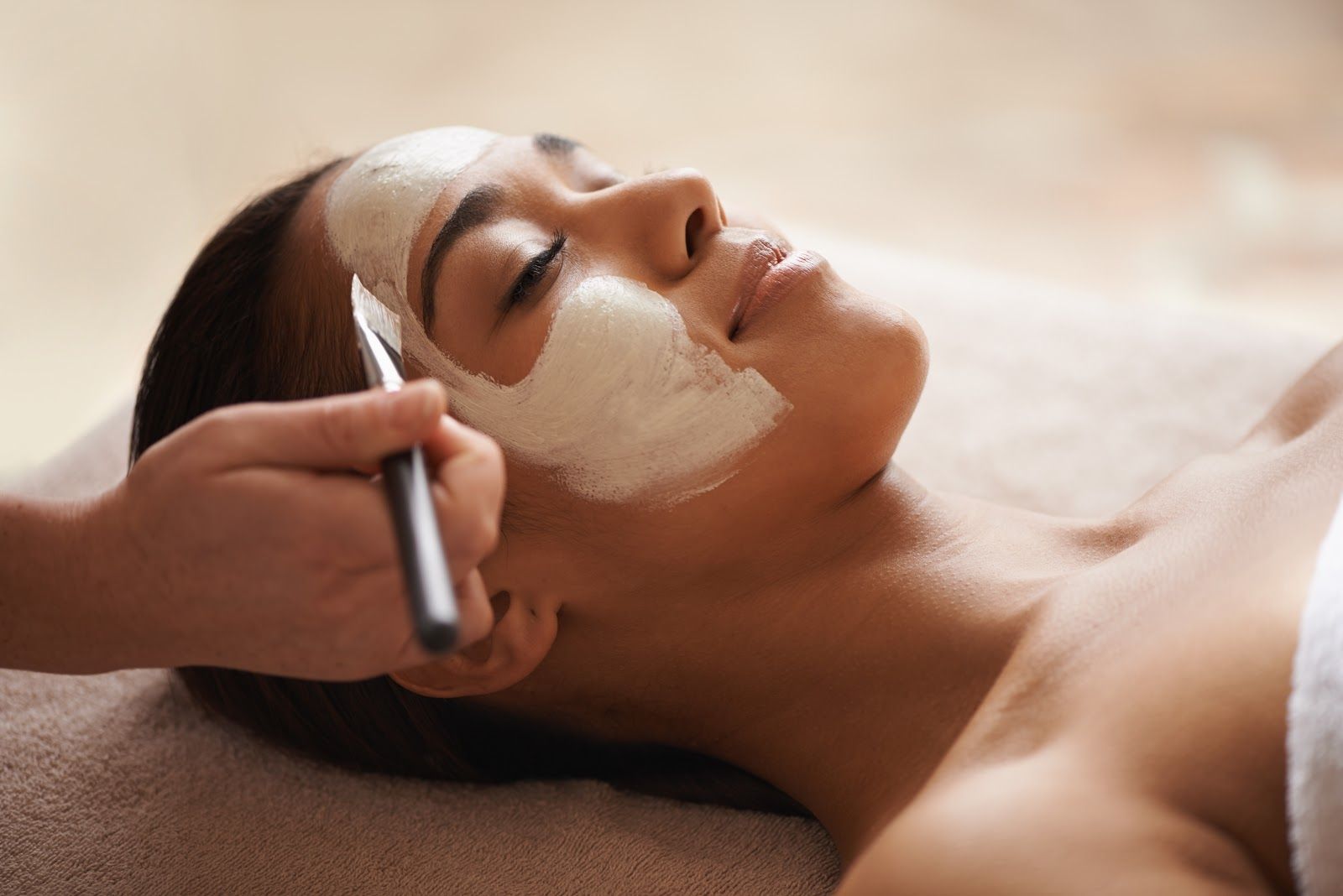Woman receiving a facial treatment while a brush applies cream to her cheek