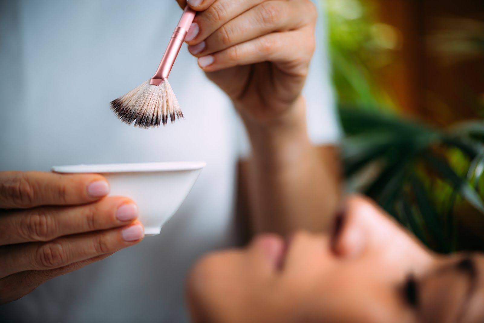 Close-up of a facial mask being applied with a brush beside a person holding a small bowl