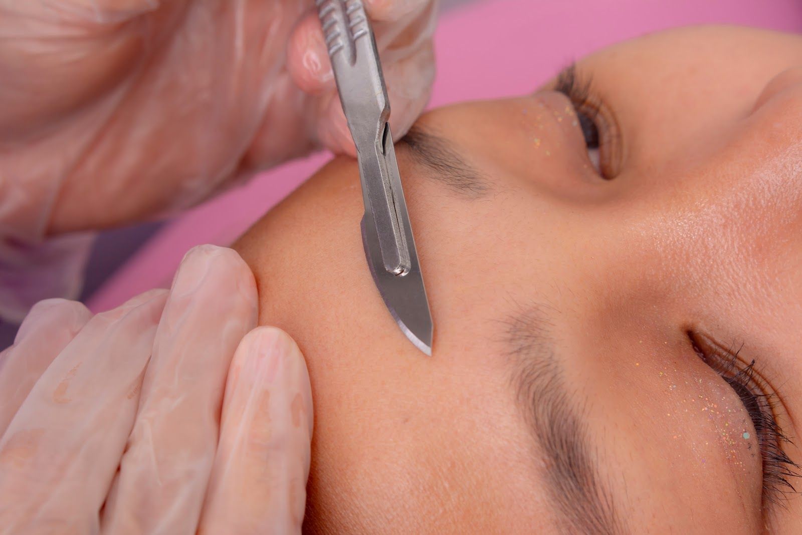 Cosmetician shaping eyebrow with a metal blade on a client's closed eye area