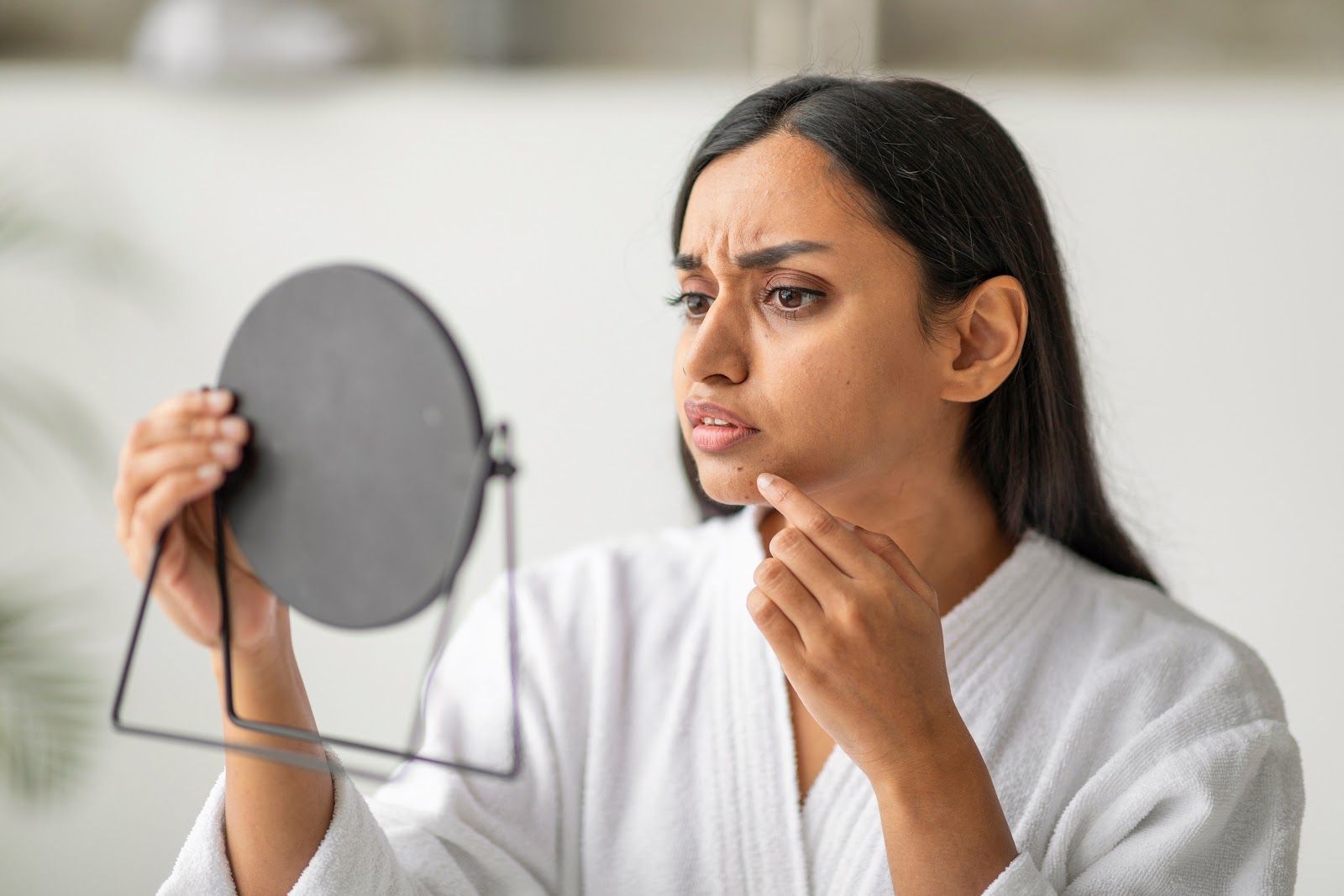 Woman in a white robe examining a mirror with a concerned expression and touching her chin.