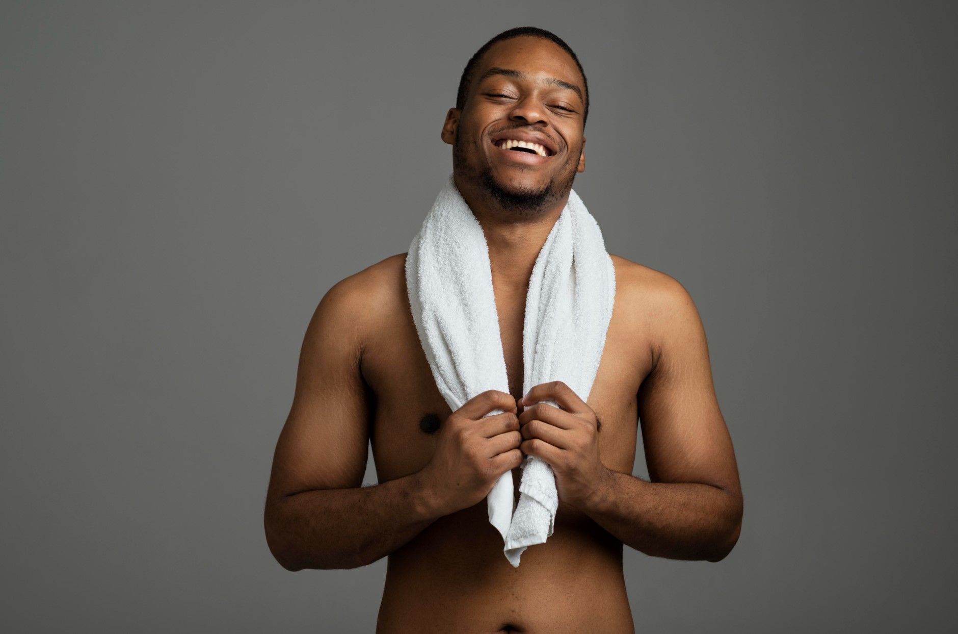 Smiling shirtless man holding a white towel around his neck against a gray background
