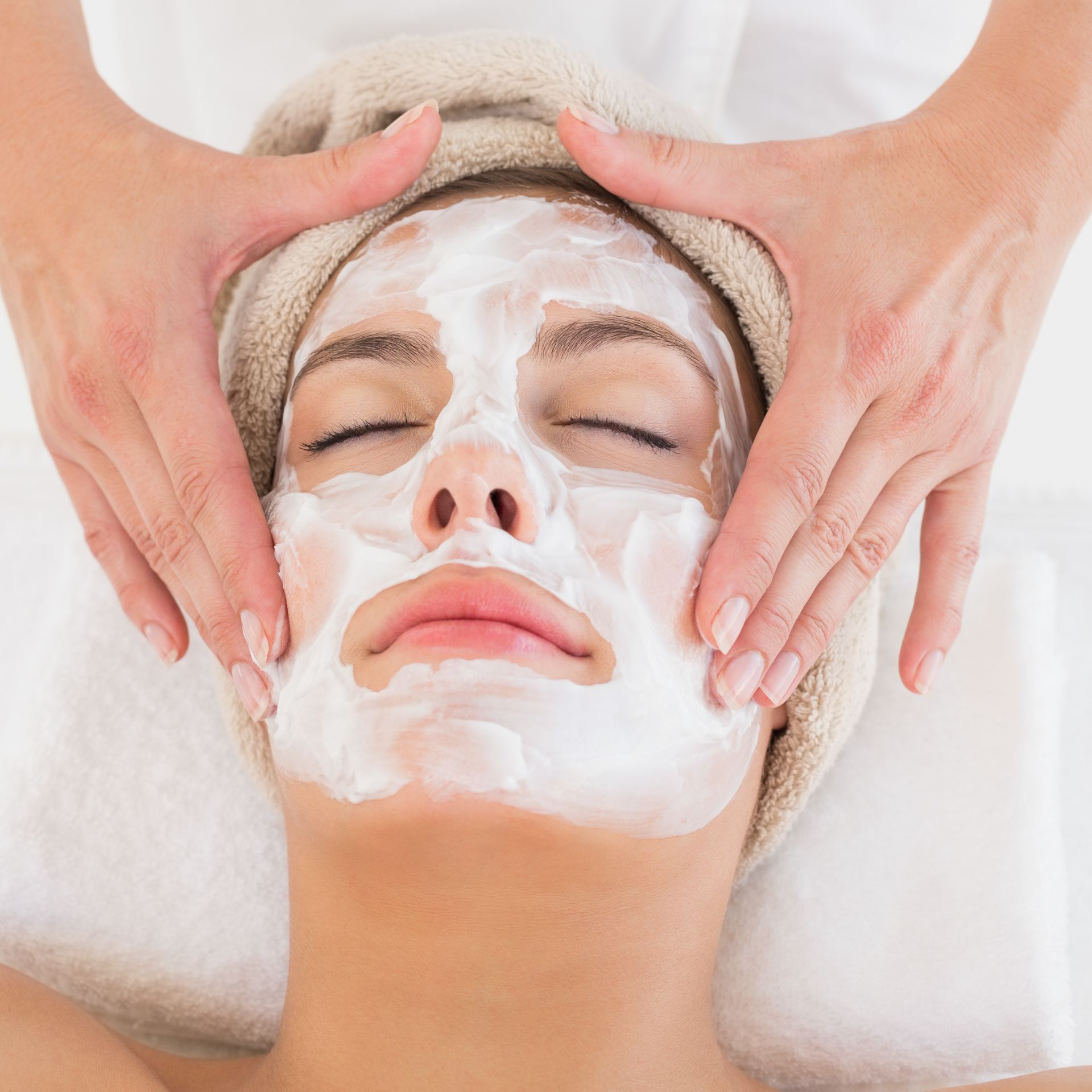 A person receiving a facial treatment with white cream applied to their face, while a technician performs a massage.