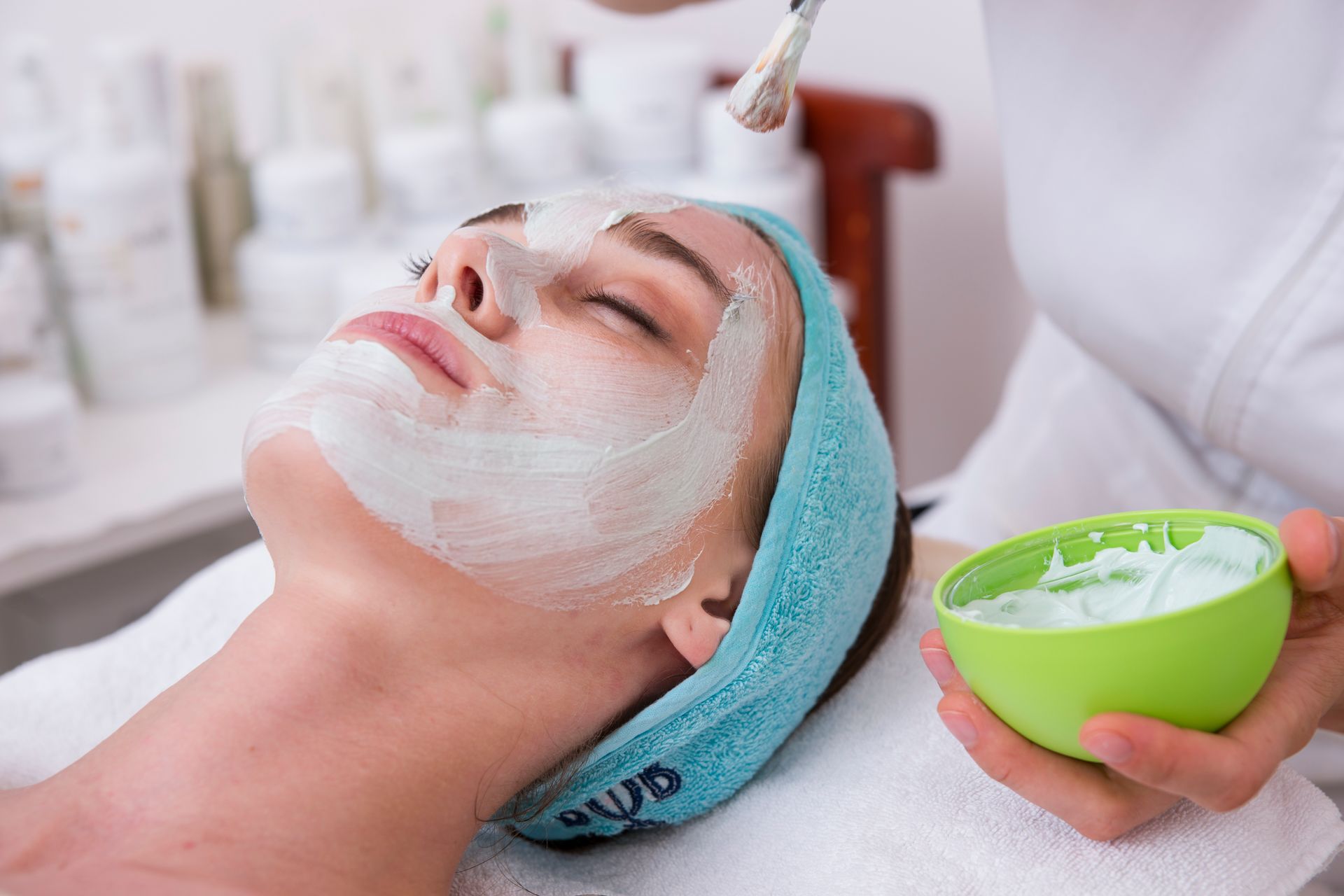 A person receiving a facial treatment with a light green cream applied to their face by an aesthetician.