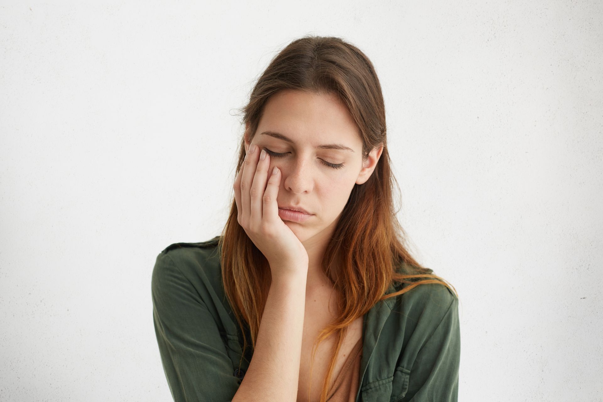 Woman with long brown hair resting her cheek on her hand, looking tired against a white wall