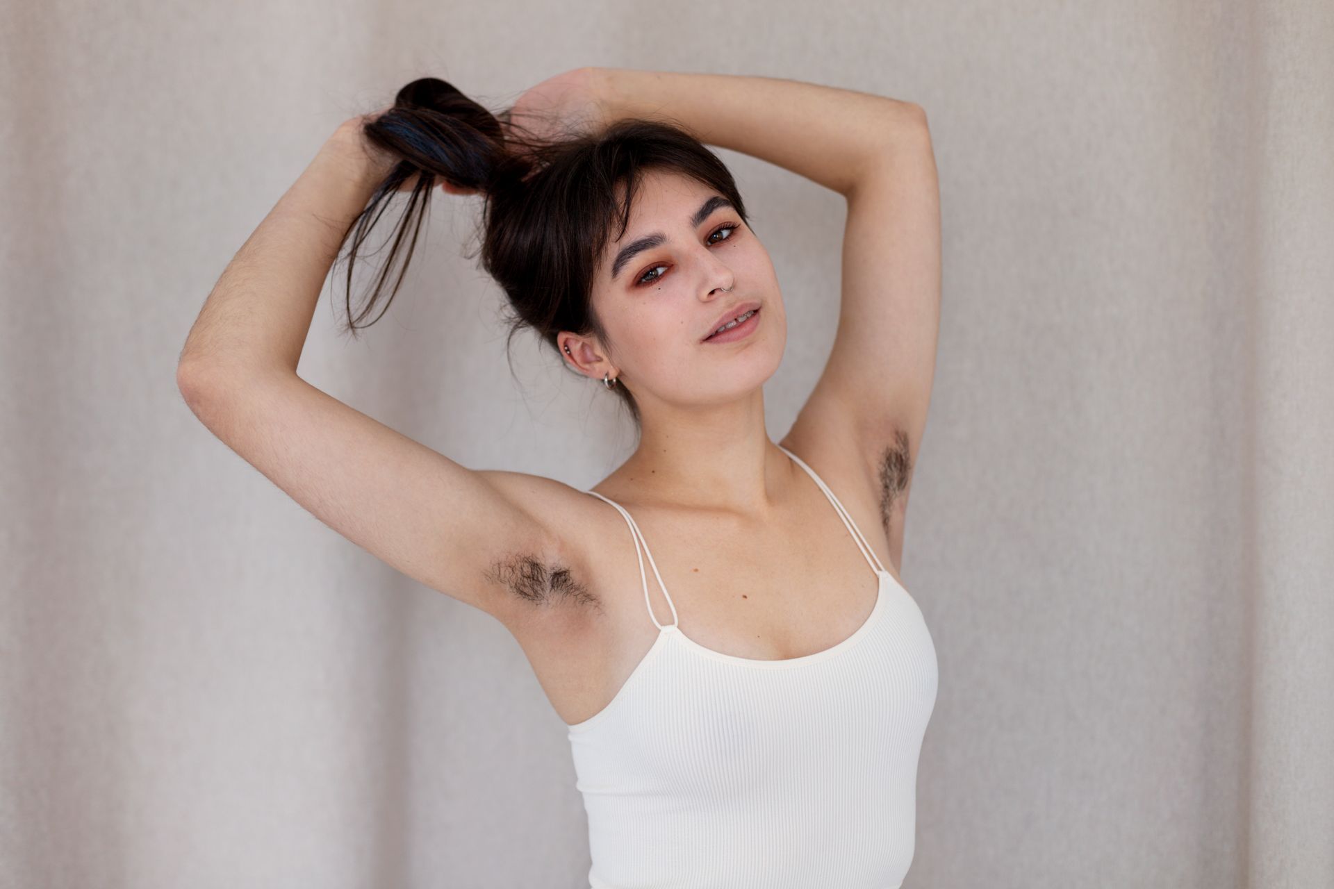 Woman in a white tank top poses with arms raised against a light curtain backdrop.