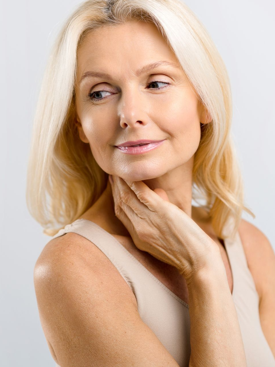 Portrait of a blonde woman in a white top gently touching her neck against a light background