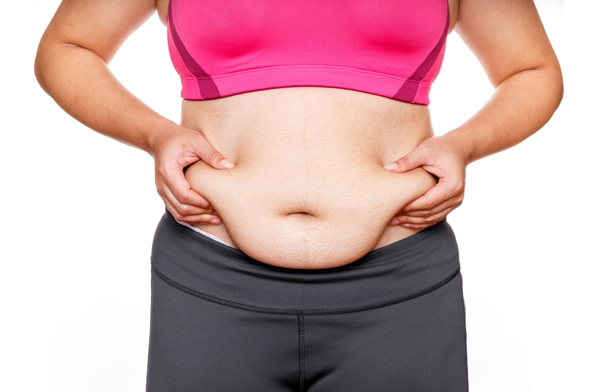 Person pinching belly fat above black leggings, wearing a pink sports bra against a white background