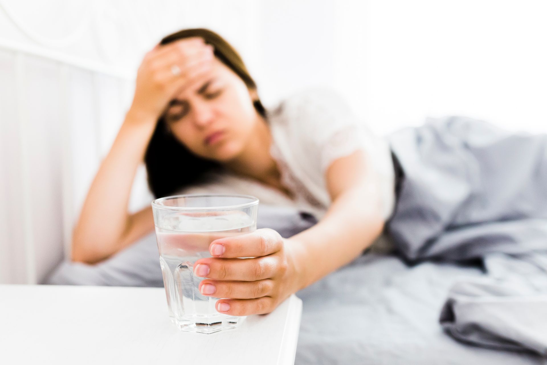 Person lying in bed, reaching for a glass of water while holding their forehead, looking unwell