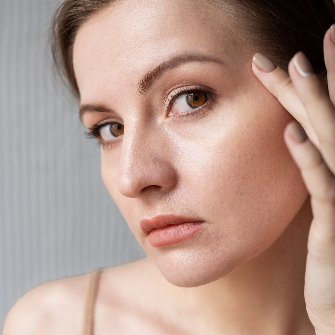 Close-up portrait of a person with a neutral expression, touching their face in soft light.
