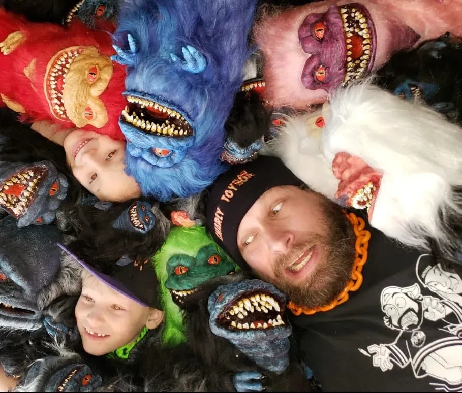 A man and two children surrounded by colorful, fuzzy monster puppets with sharp, detailed teeth.