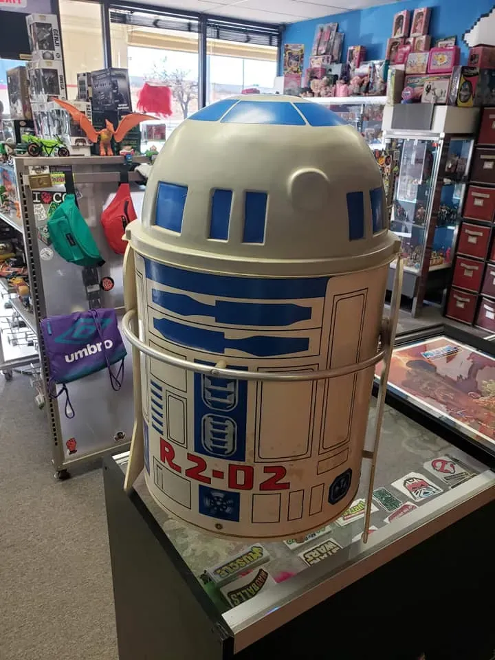 A vintage R2-D2 trash can with blue and white details, sitting on a store counter filled with various merchandise.