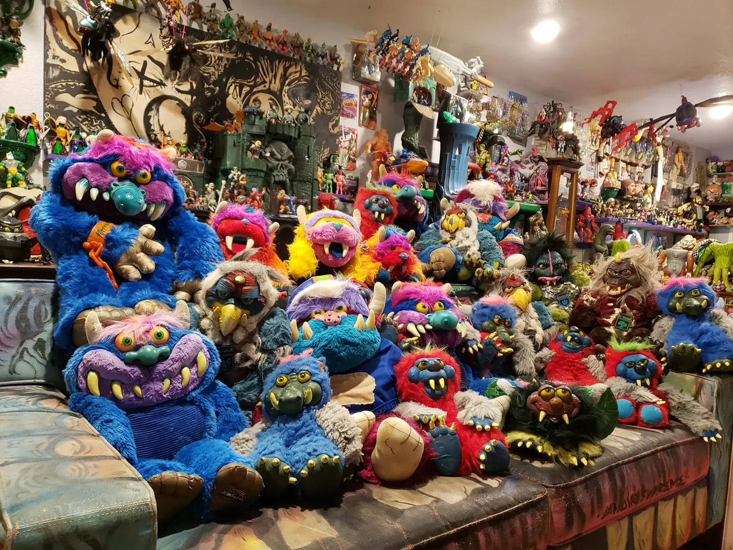 A large collection of colorful, furry monster dolls with wide grins, gathered on a couch in a room full of toys. Featuring My Pet Monsters, My pet monster Pets, Los Temblors, Defensors De La Neutrlaza and more!