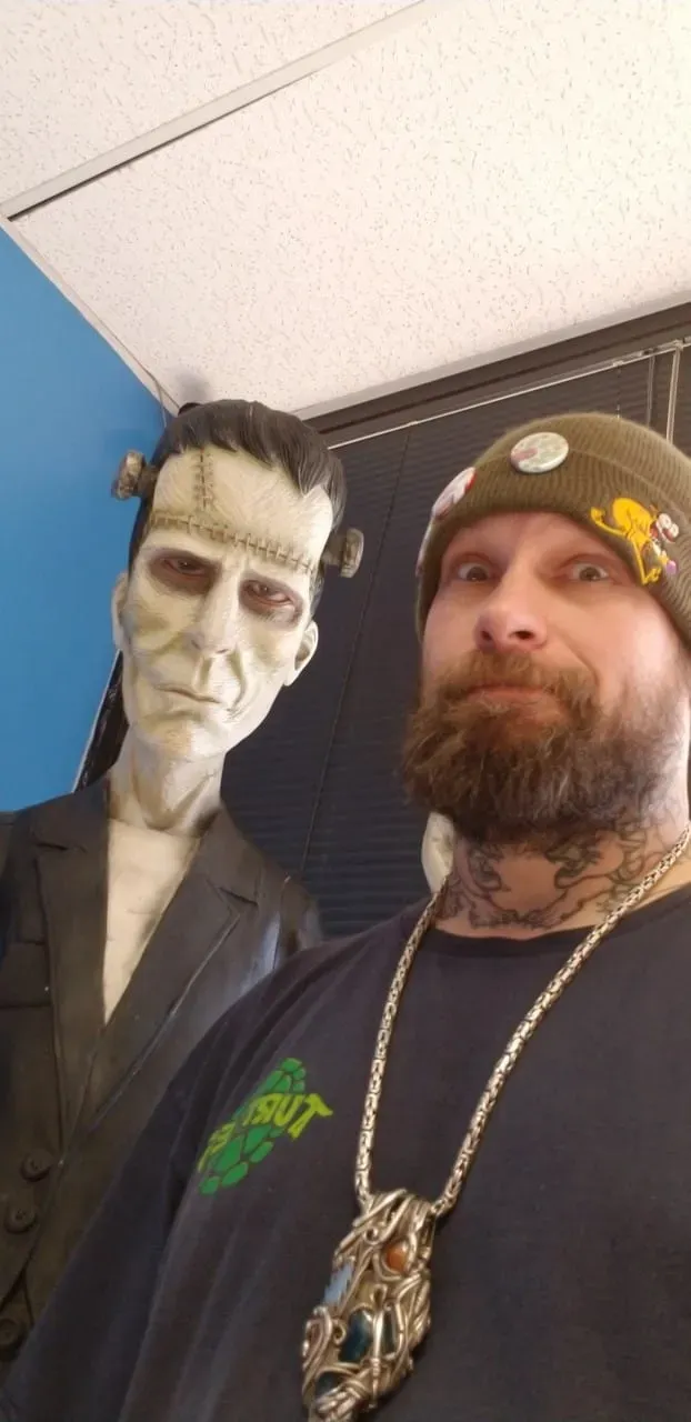 A person with a beard and tattoos wearing a beanie poses next to a life-sized statue of Frankenstein's monster.