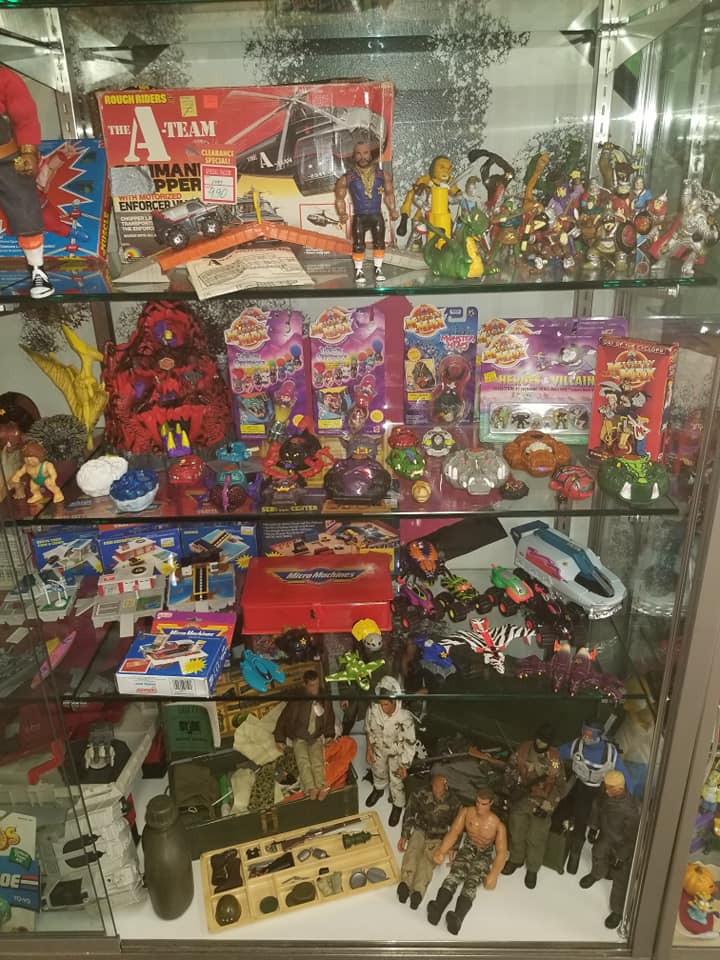A glass display case filled with various vintage action figures, toy vehicles, and boxed collectibles on multiple shelves.