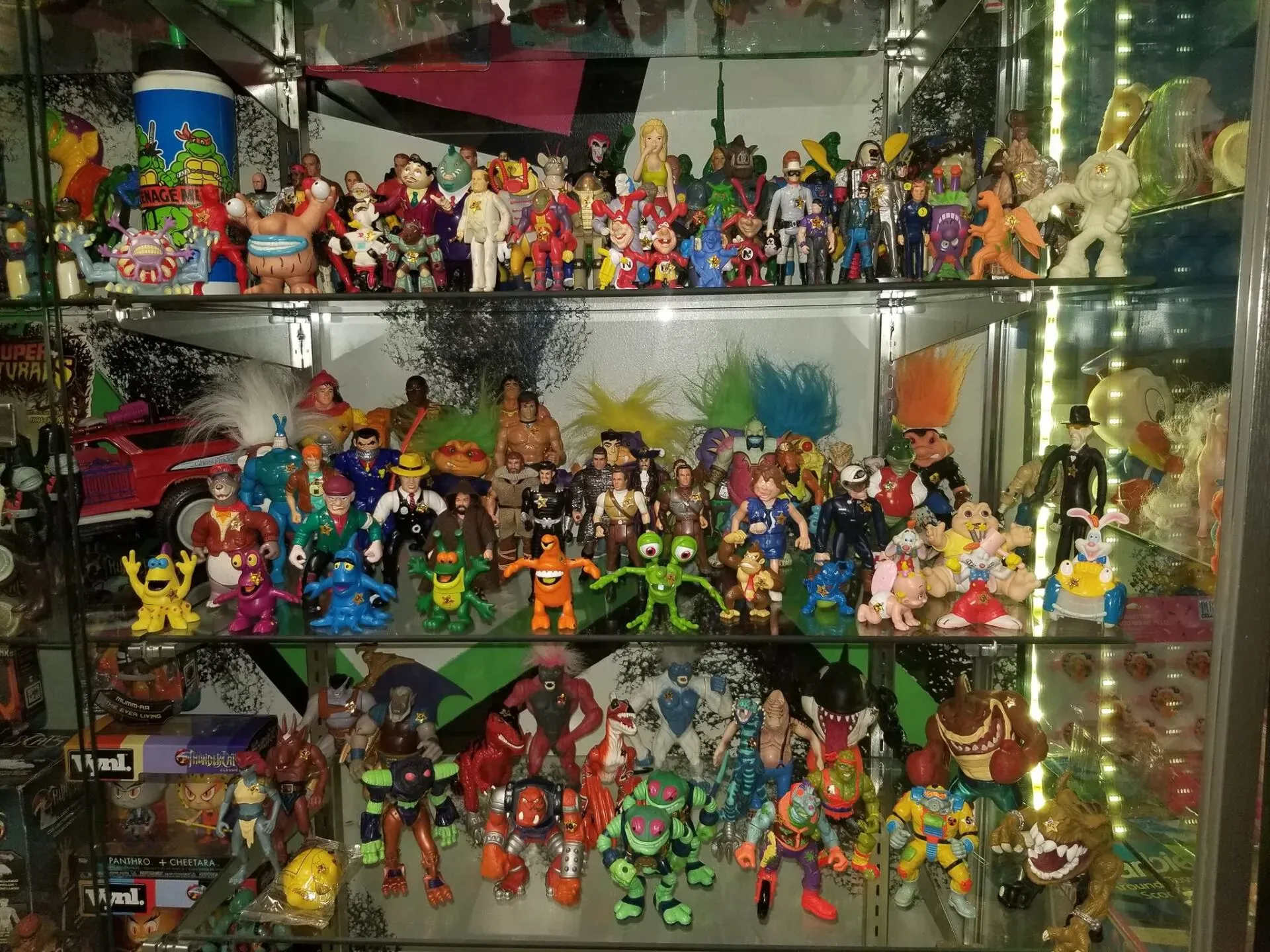 A multi-level glass display case filled with a large collection of colorful, vintage action figures and toys.