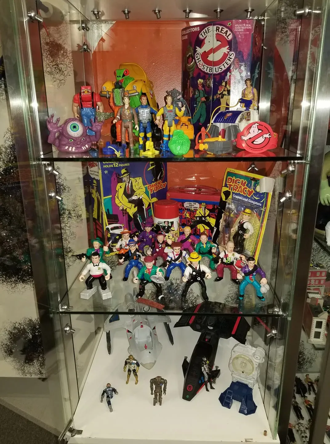 A glass display case featuring three shelves filled with vintage action figures, including Ghostbusters and Batman items.