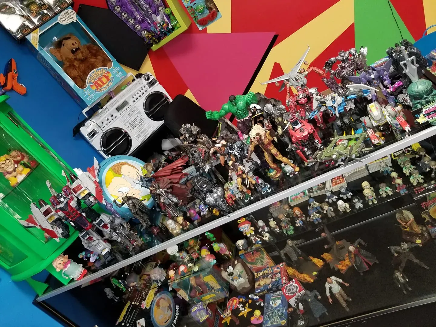 A collection of action figures, collectibles, and a boombox displayed on shelves against a colorful geometric wall.