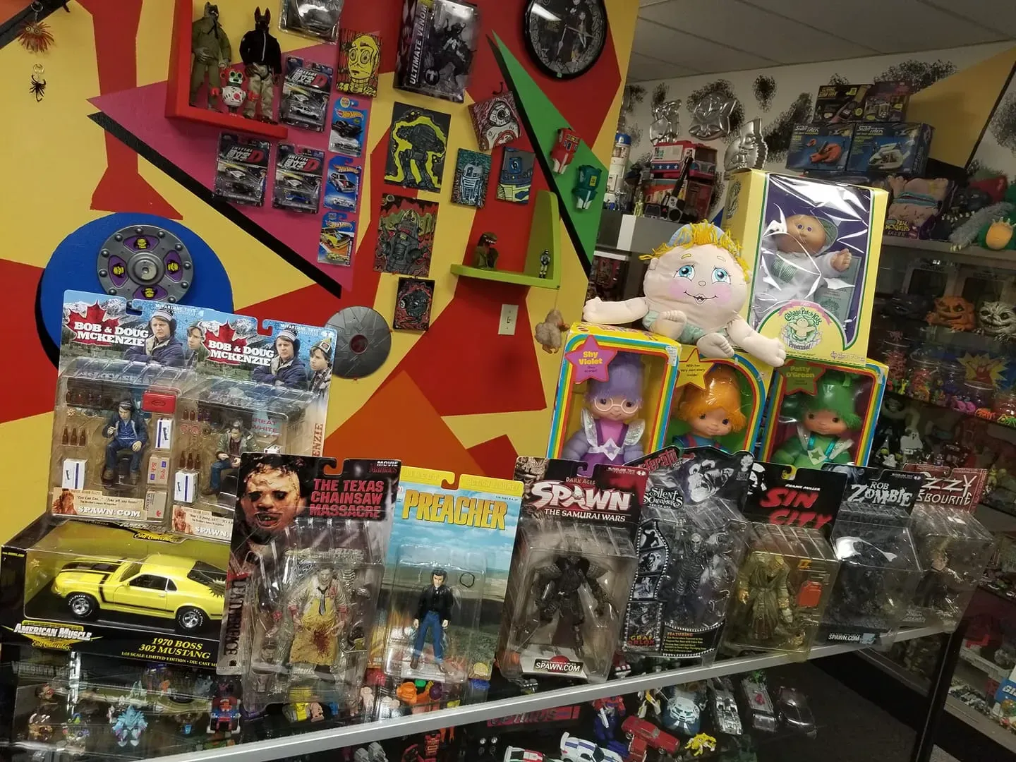 A retail display of vintage action figures, dolls, and toy collectibles on shelves against a geometric, colorful wall.
