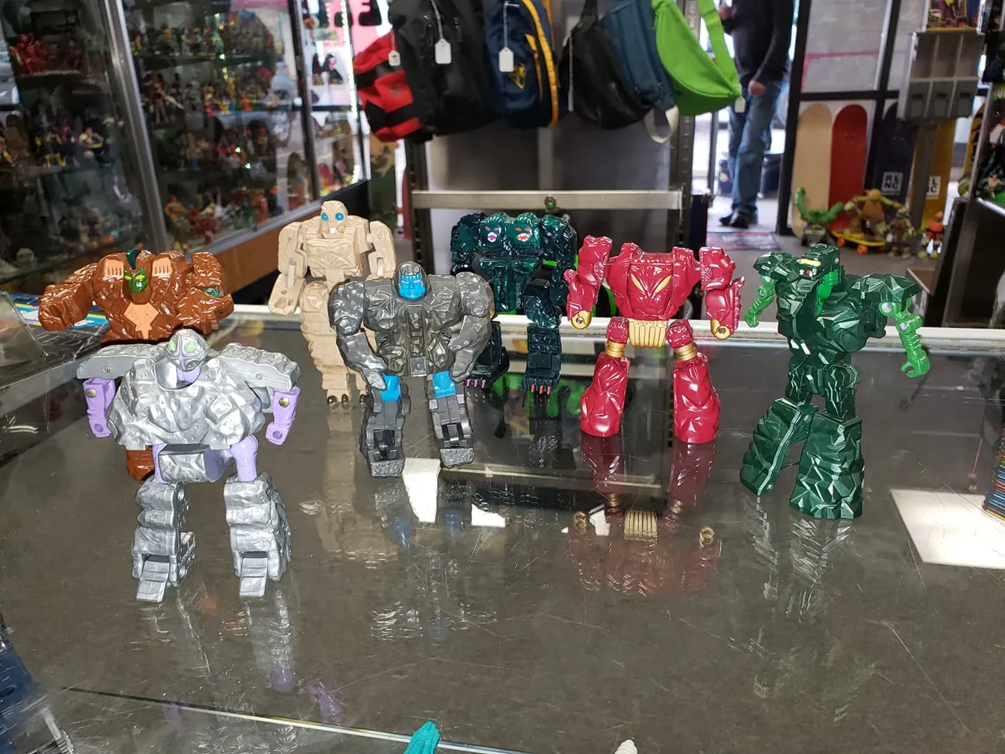 Seven colorful, blocky robot action figures stand on a glass display counter in a store filled with toys.