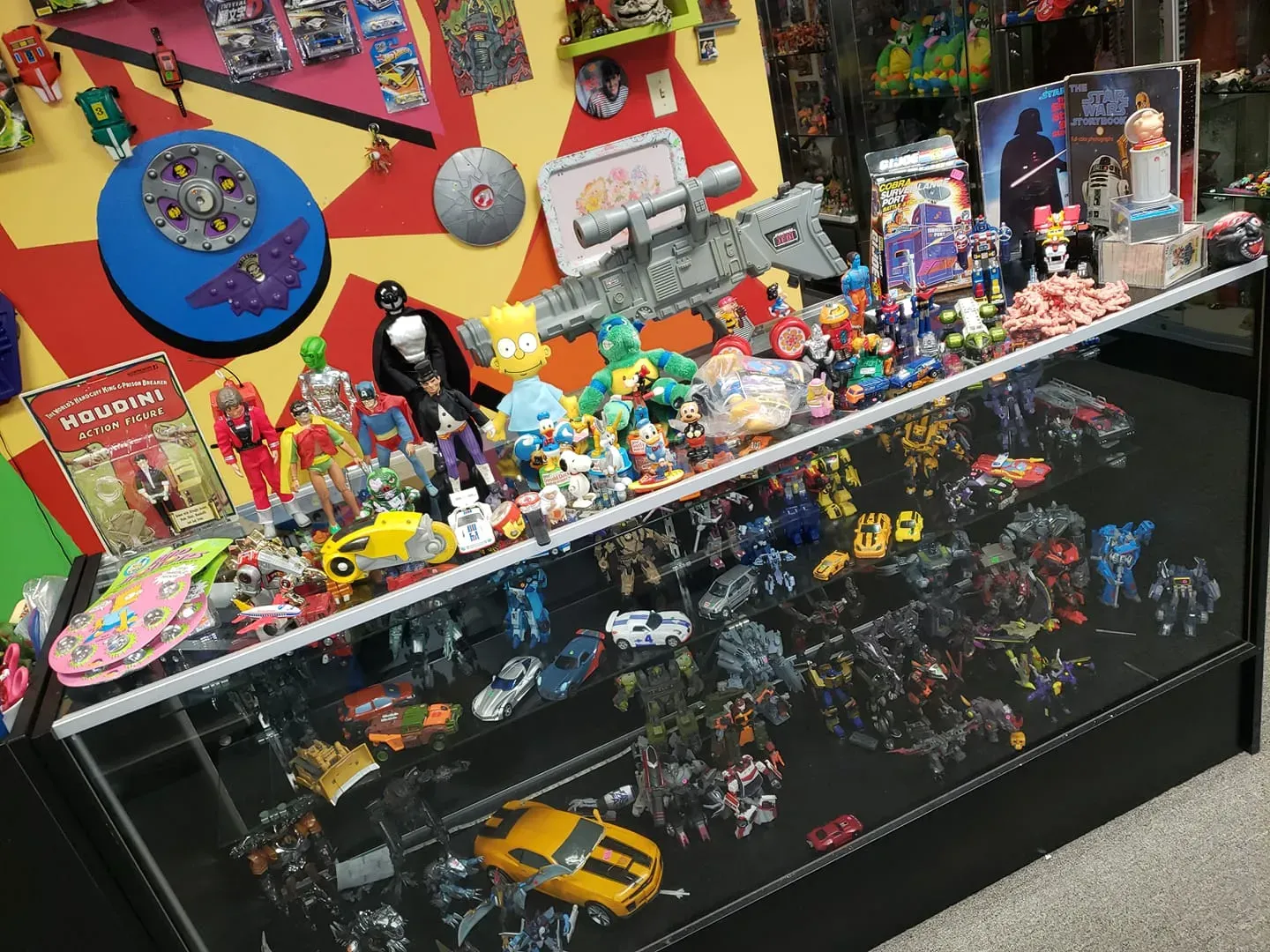 A glass display case filled with various toys, figures, and collectibles, set against a colorful, geometric-patterned wall.