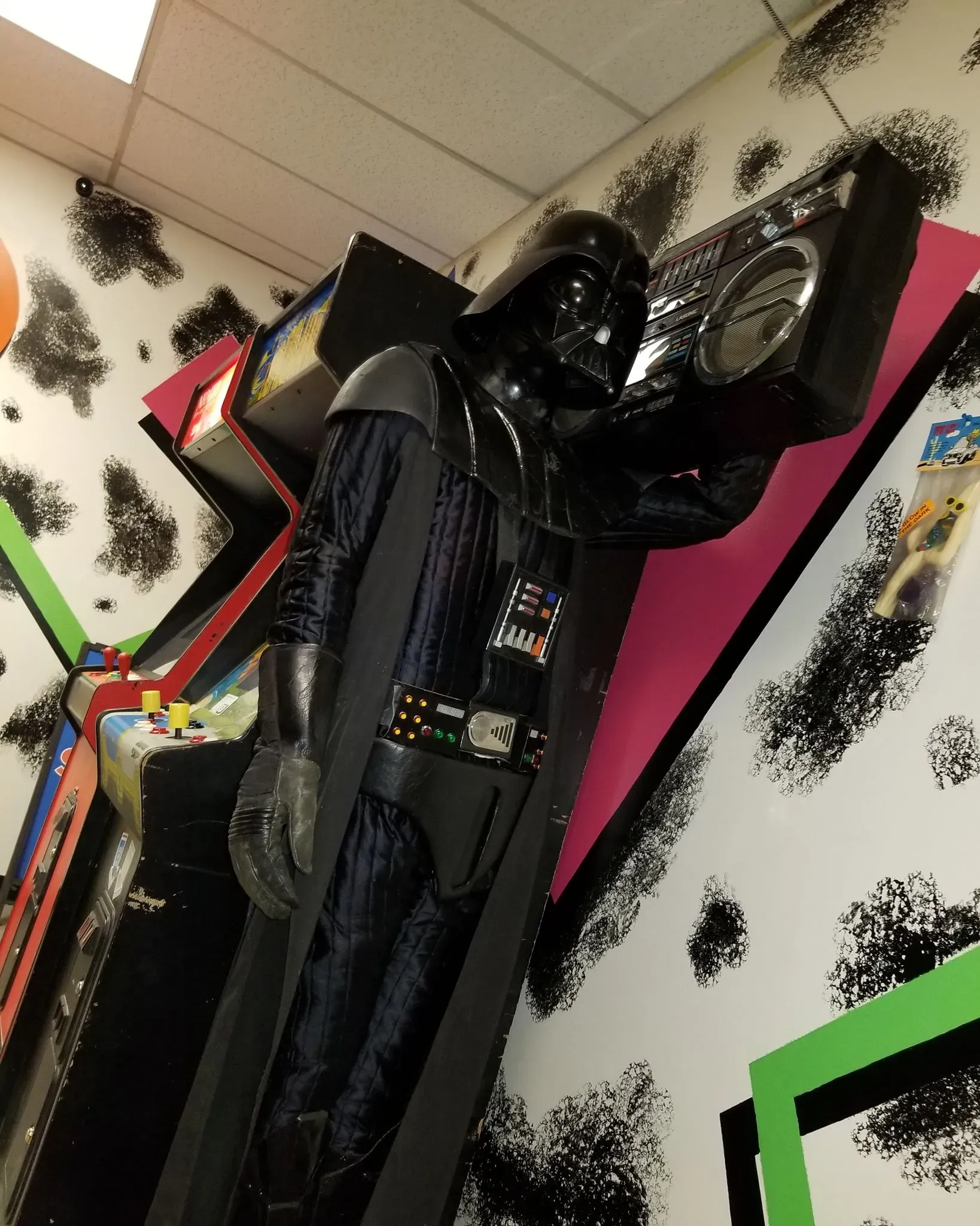 A Darth Vader statue stands in an arcade-themed room, holding a boombox over its shoulder.