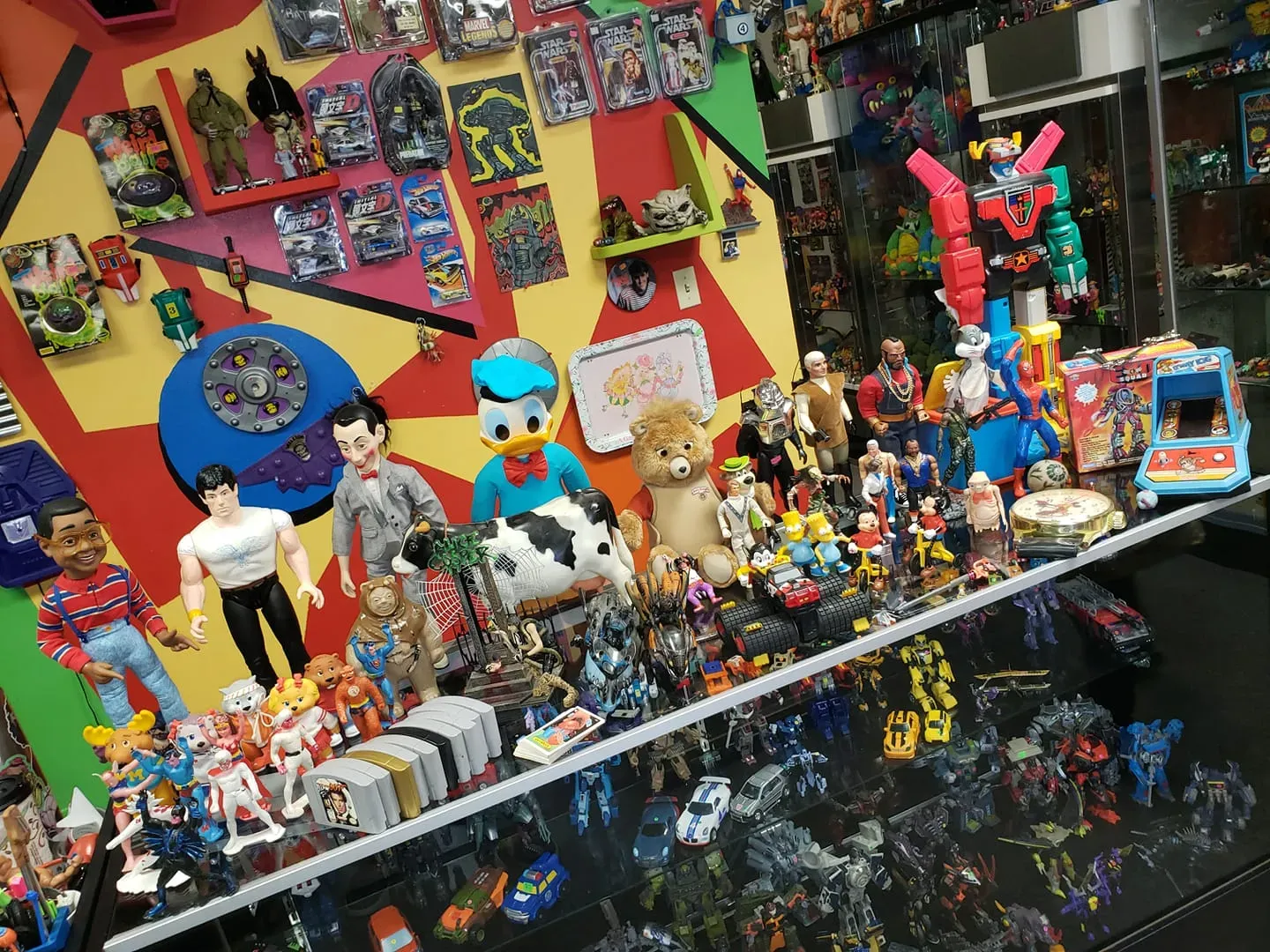 A colorful display shelf filled with vintage action figures, dolls, and toys, set against a geometric patterned wall.