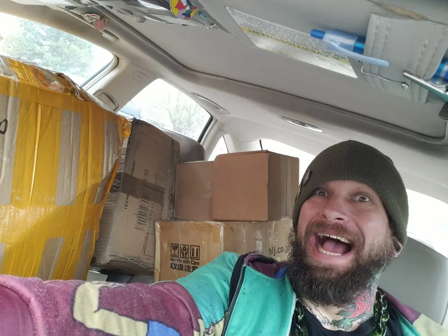 A bearded person wearing a beanie and colorful clothing smiles excitedly in a car packed with cardboard shipping boxes.