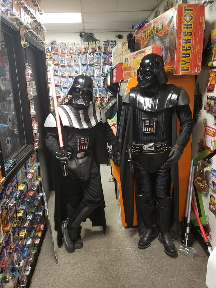 Two individuals dressed in full Darth Vader costumes stand in a toy store aisle lined with many packaged toys.