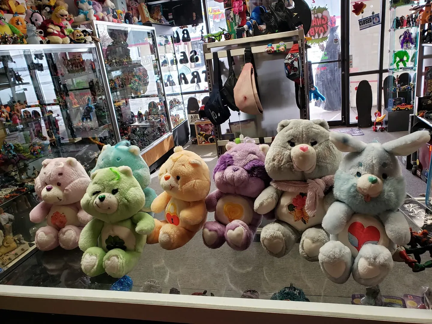 A row of six plush Care Bear-style toys in various colors and designs sitting on a glass display case in a shop.