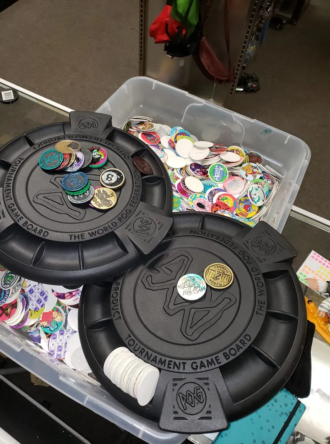 Two black plastic POG tournament game boards sit atop a clear storage bin filled with a collection of colorful POGs.