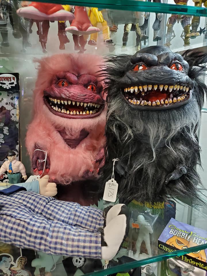 Two furry, sharp-toothed Critters puppets, one pink and one dark gray, displayed inside a glass store shelf.
