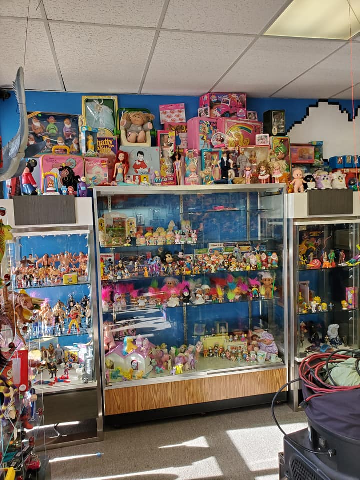 A shop filled with shelves and glass display cases stocked with various vintage toys and dolls against a blue wall.