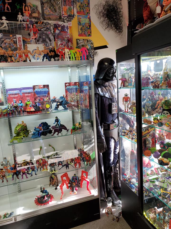 A large Darth Vader figure stands between two glass display cases filled with various action figures and collectibles.