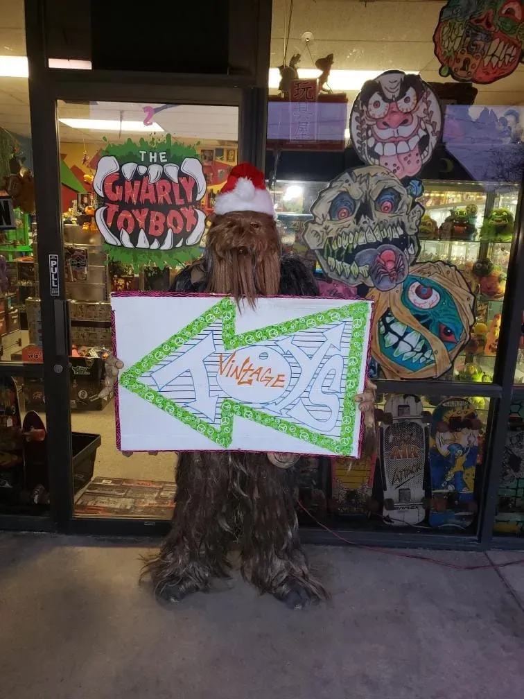 A person in a Chewbacca costume wearing a Santa hat holds a green-outlined sign pointing left outside