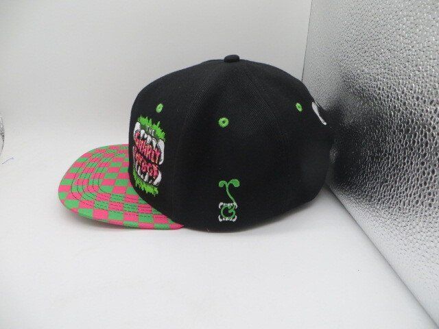 A black baseball cap with a green and pink checkered brim and a graphic of monster teeth on the side.