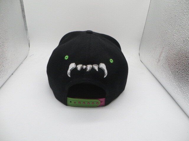 A black snapback cap featuring an embroidered white monster mouth with teeth and small green dots on a white background.