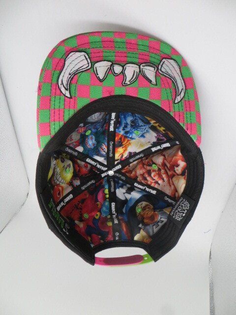 A flat-brimmed cap with a pink and green checkerboard pattern featuring a graphic of monster teeth on the underside.