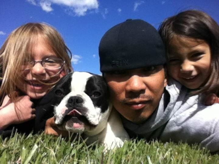 Man, two girls, and Boston Terrier puppy smiling on grass under blue sky.