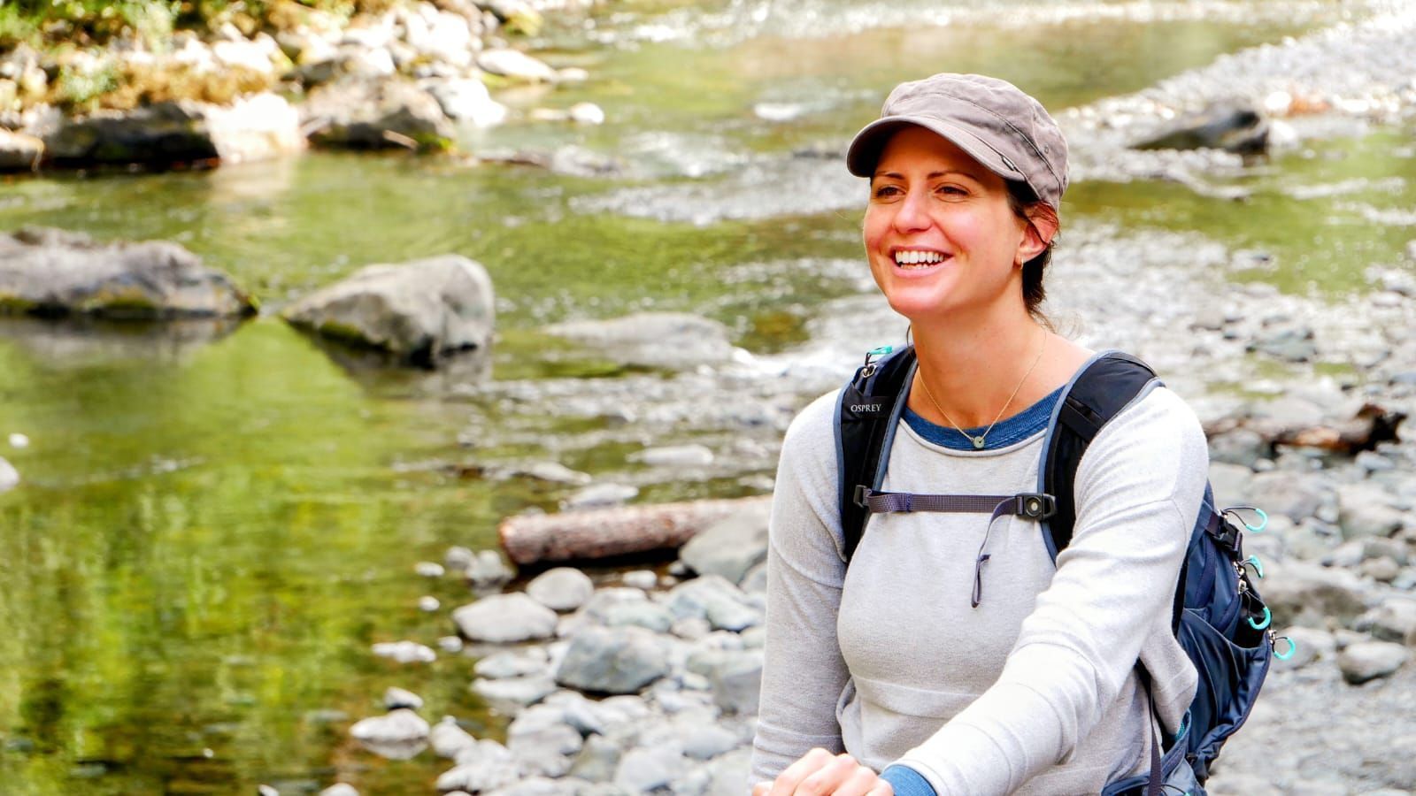 Smiling hiker in a gray cap and backpack sitting by a rocky stream in a green forest