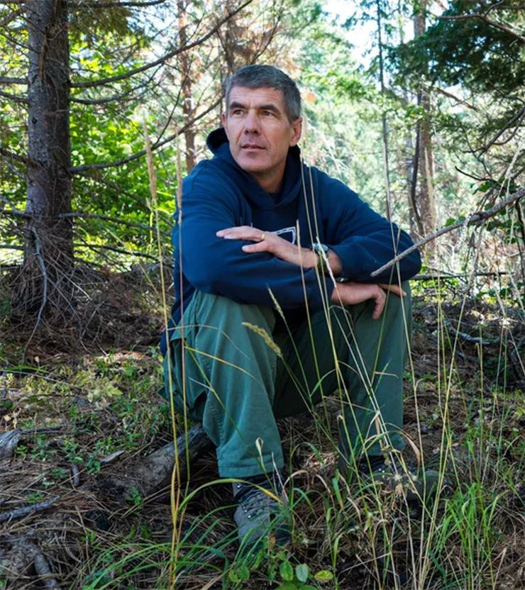 Man crouching in a forest among trees and tall grass, wearing a navy jacket and green pants