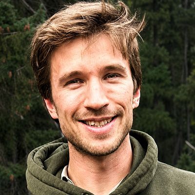 Smiling person in an olive green hoodie outdoors with trees in the background