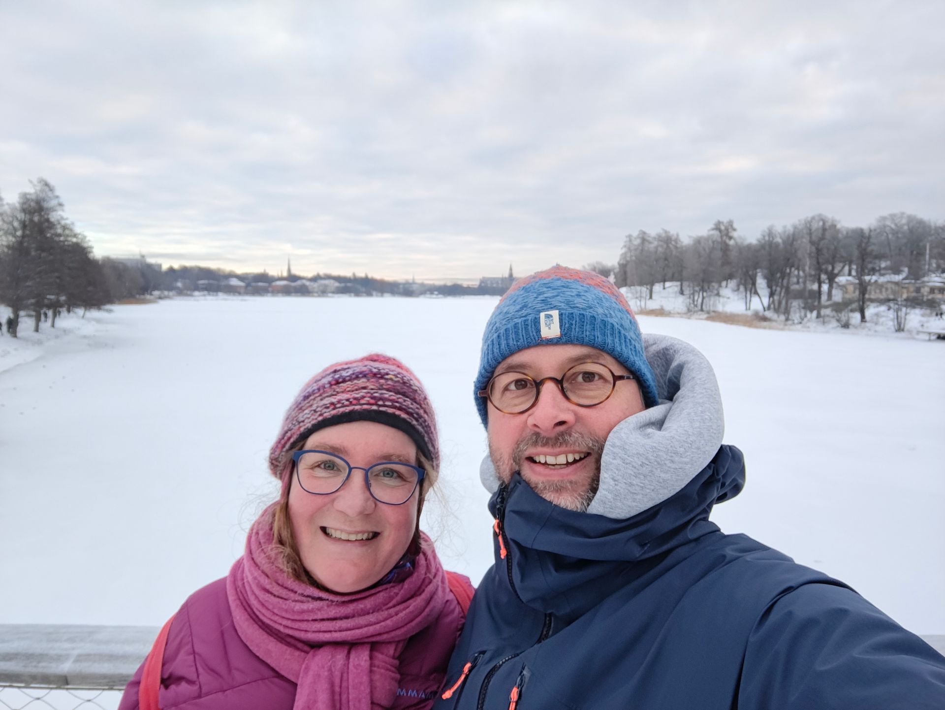 Twee mensen glimlachen met wintermutsen en sjaals op een besneeuwde rivieroever onder een bewolkte hemel.