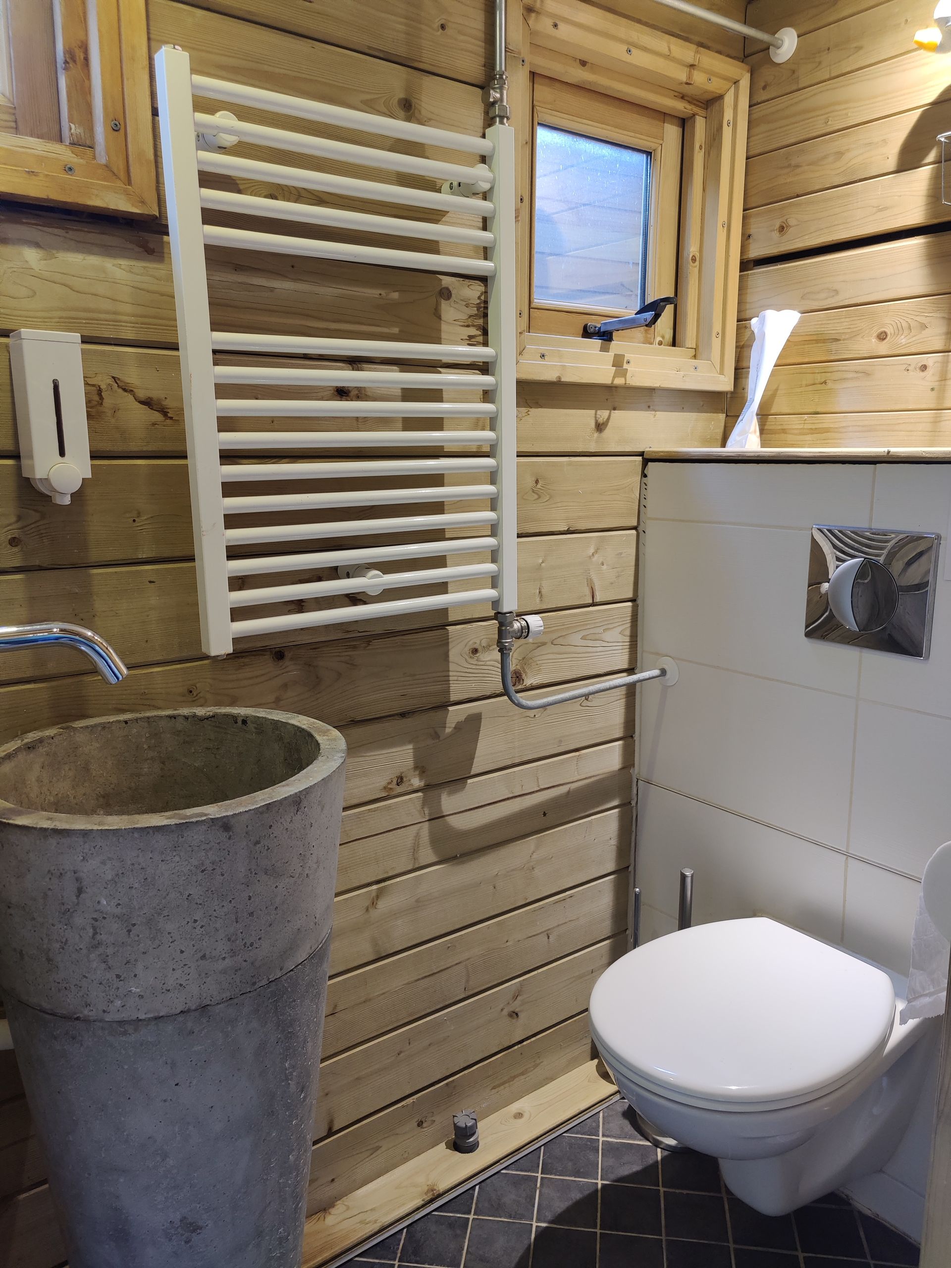 Kleine badkamer met houten lambrisering, toilet, betonnen wastafel, witte handdoekradiator en een klein raam.