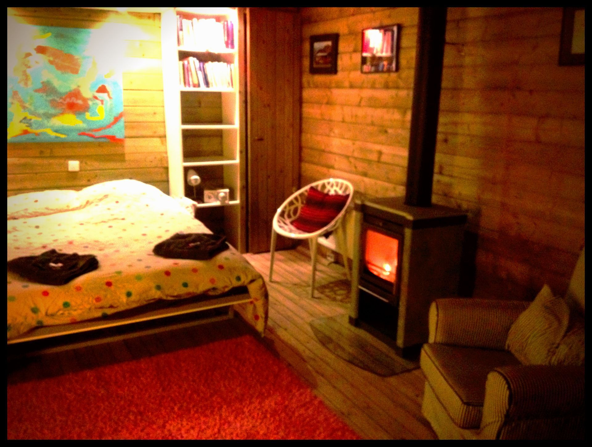 Gezellige slaapkamer in een blokhut met bed, rood tapijt, stoel en een gloeiende houtkachel naast een bakstenen muur.