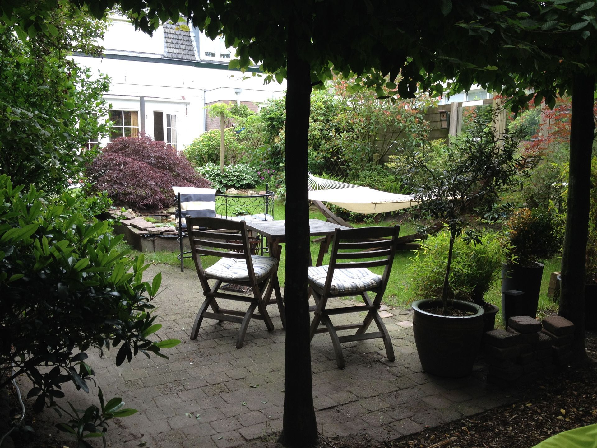 Tuinpatio met houten stoelen en tafel onder een schaduwrijke pergola, omgeven door weelderig groen.