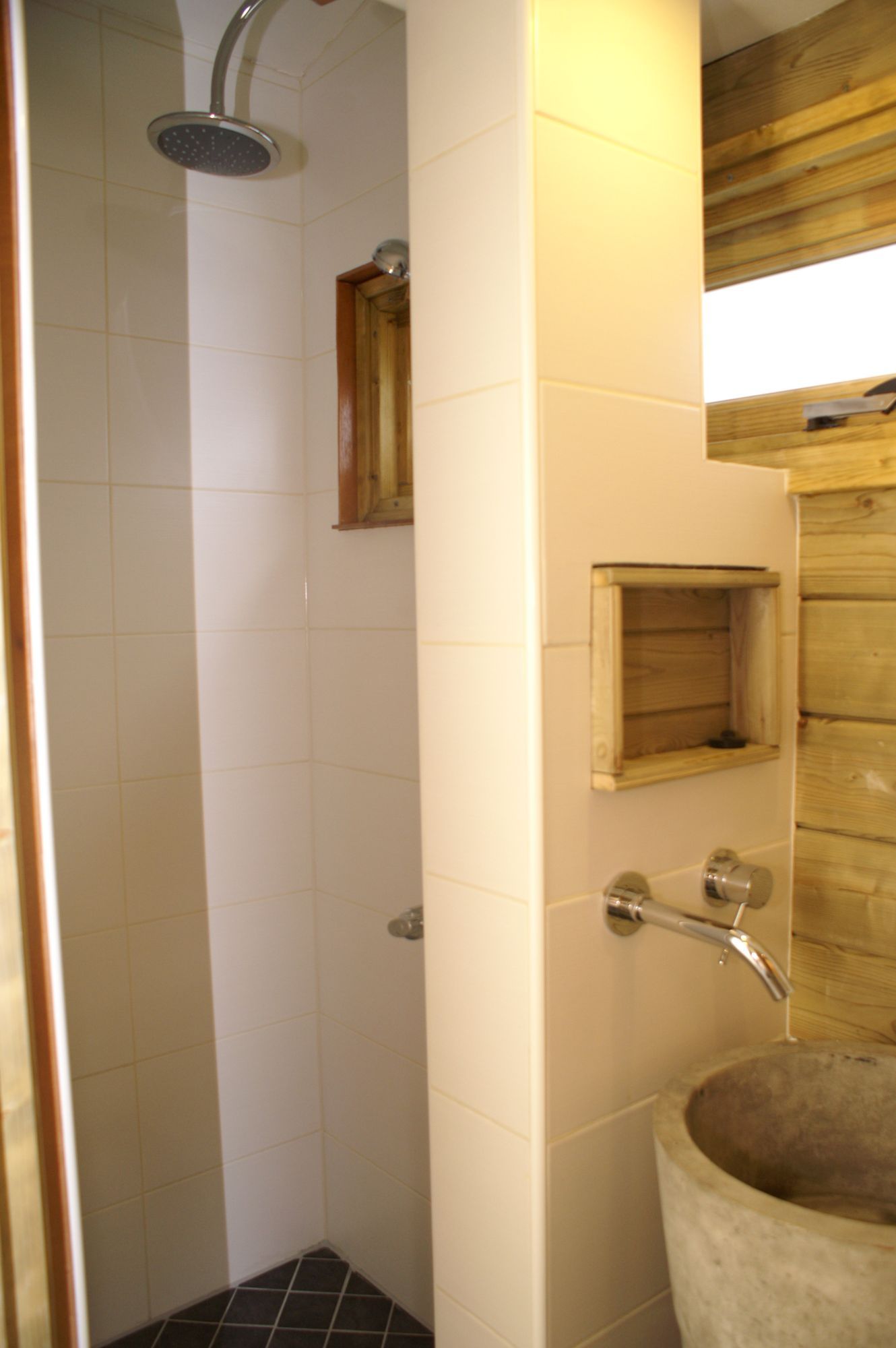 Kleine betegelde douche- en wastafelruimte met chromen douchekop en kraan, plus houten raamjaloezieën.