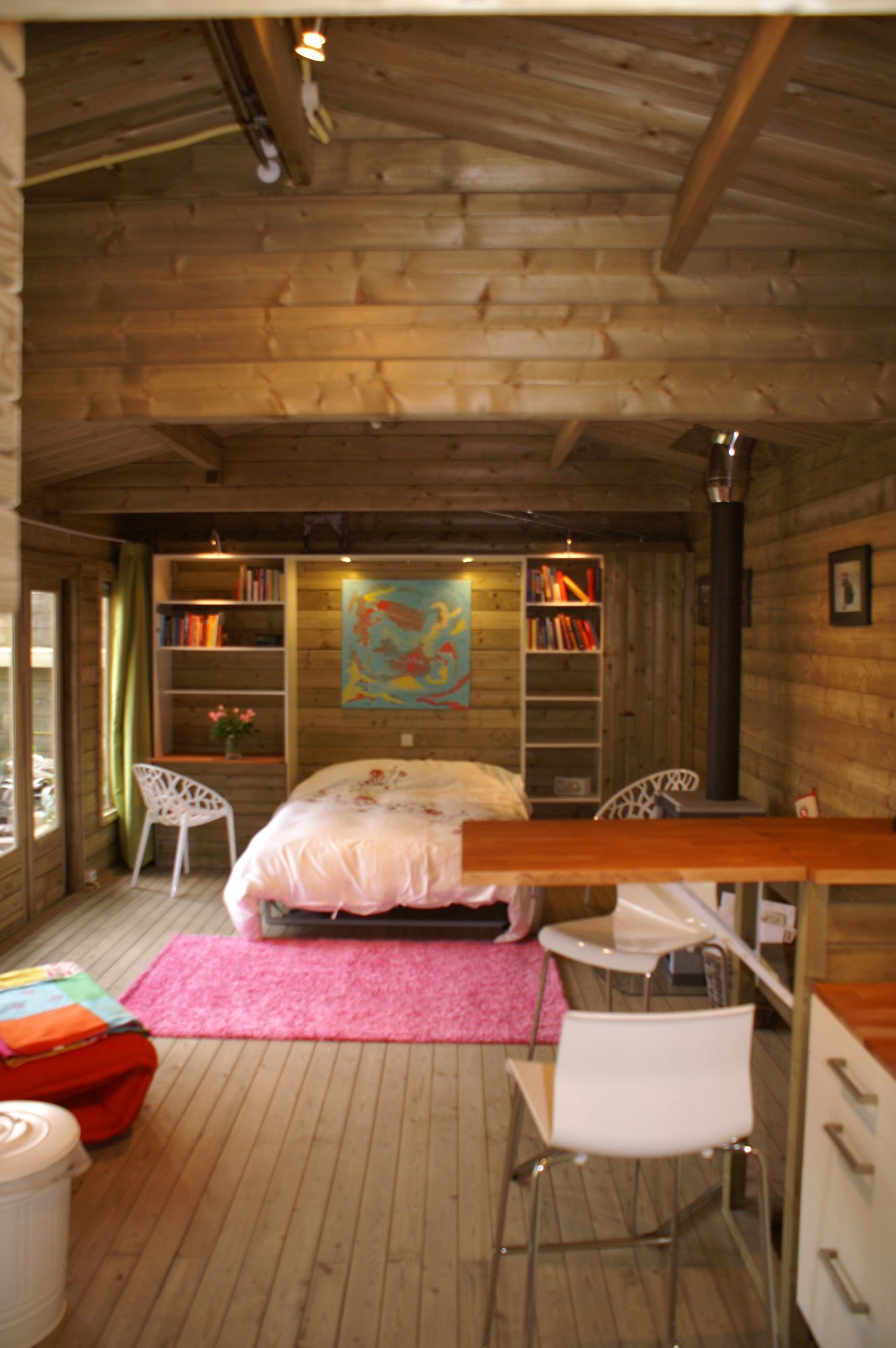 Rustieke slaapkamer in een blokhut met een bed, roze vloerkleed, houten wanden, planken en een tafel op de voorgrond.