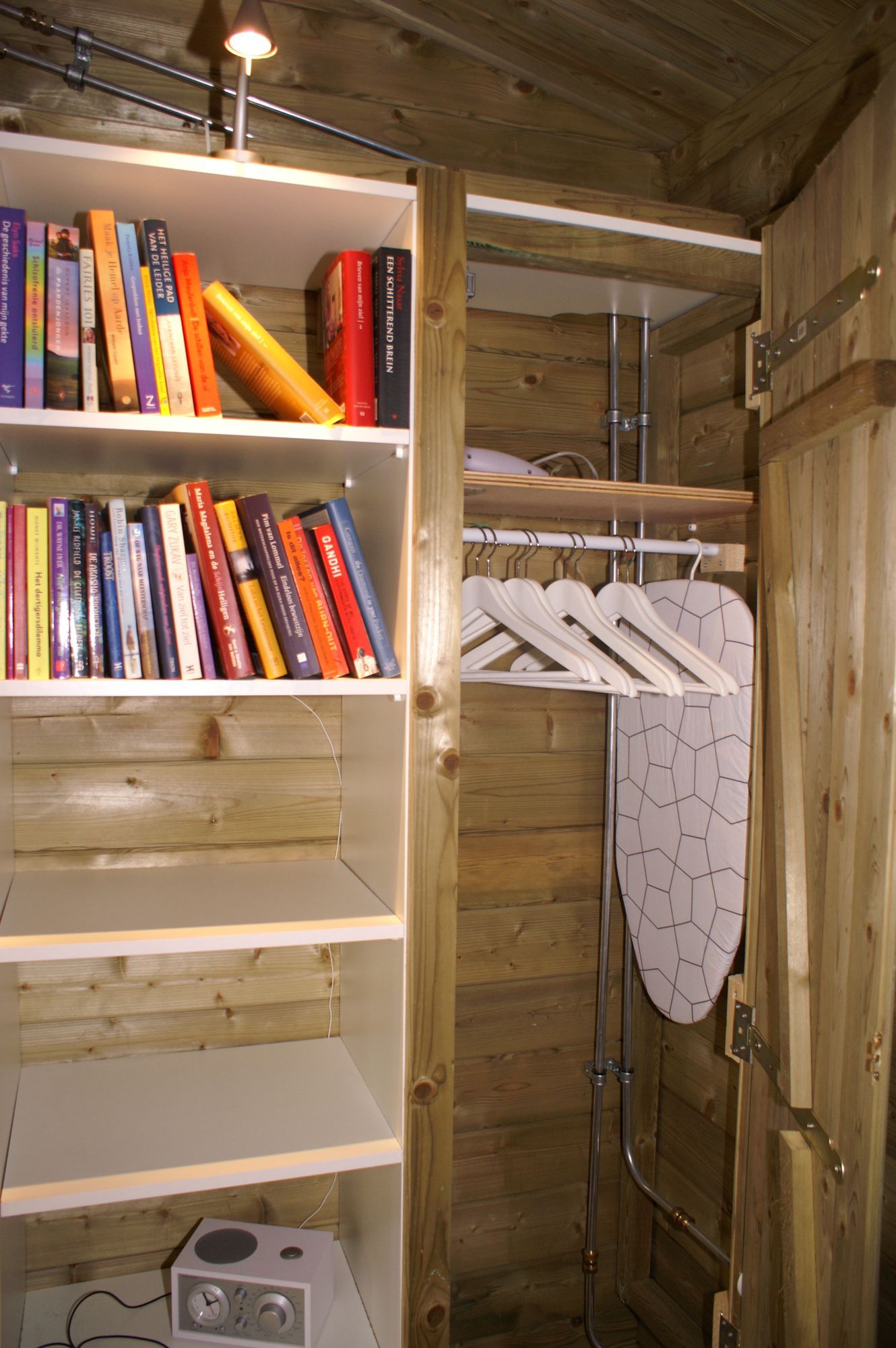 Houten kast met boeken op planken en een hangrek met witte overhemden naast een strijkplank.